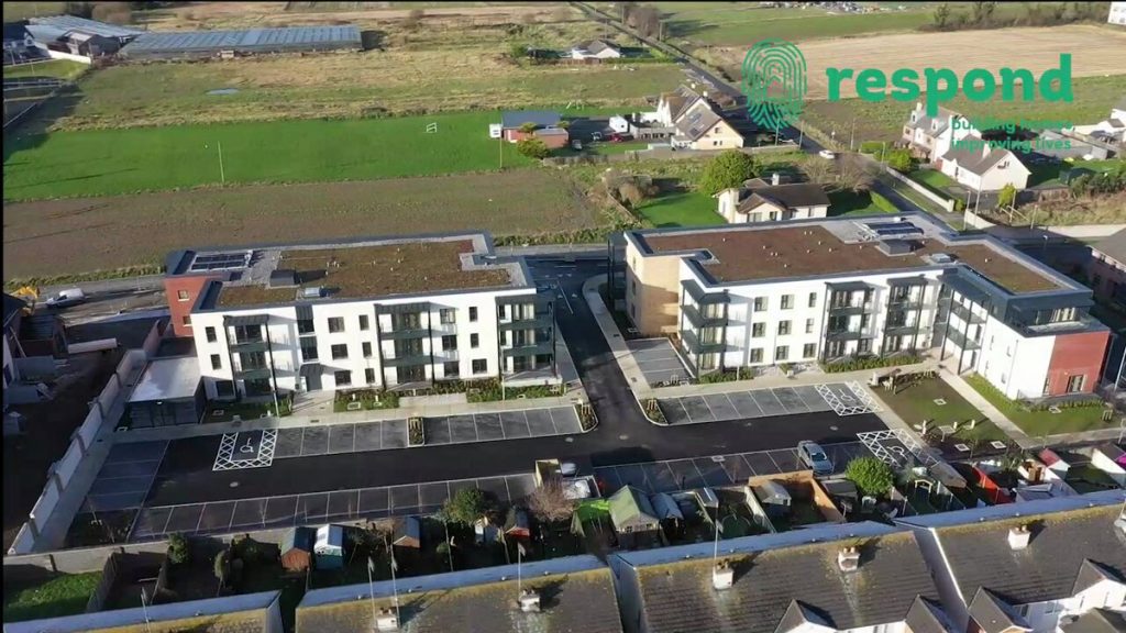 Image of an aerial view of a residential building with parking spaces in front and green fields at the back.