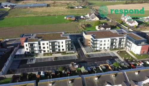 Image of an aerial view of a residential building with parking spaces in front and green fields at the back.